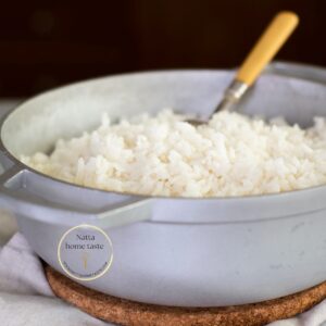 Olla de aluminio con arroz blanco cocido y esponjoso. Se observa una cuchara de mango amarillo dentro de la olla. La olla está sobre un salvamanteles de corcho y un paño blanco, con un fondo oscuro desenfocado.