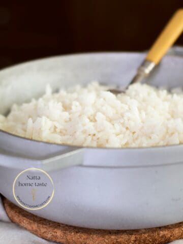 Olla de aluminio con arroz blanco cocido y esponjoso. Se observa una cuchara de mango amarillo dentro de la olla. La olla está sobre un salvamanteles de corcho y un paño blanco, con un fondo oscuro desenfocado.