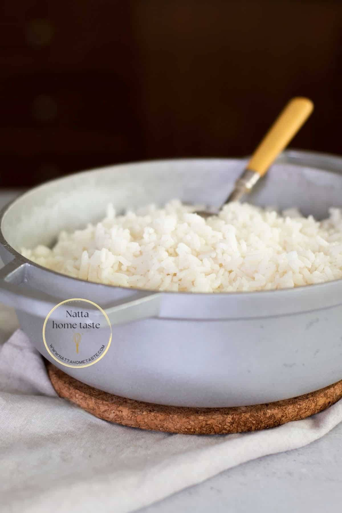 Olla de aluminio con arroz blanco cocido y esponjoso. Se observa una cuchara de mango amarillo dentro de la olla. La olla está sobre un salvamanteles de corcho y un paño blanco, con un fondo oscuro desenfocado
