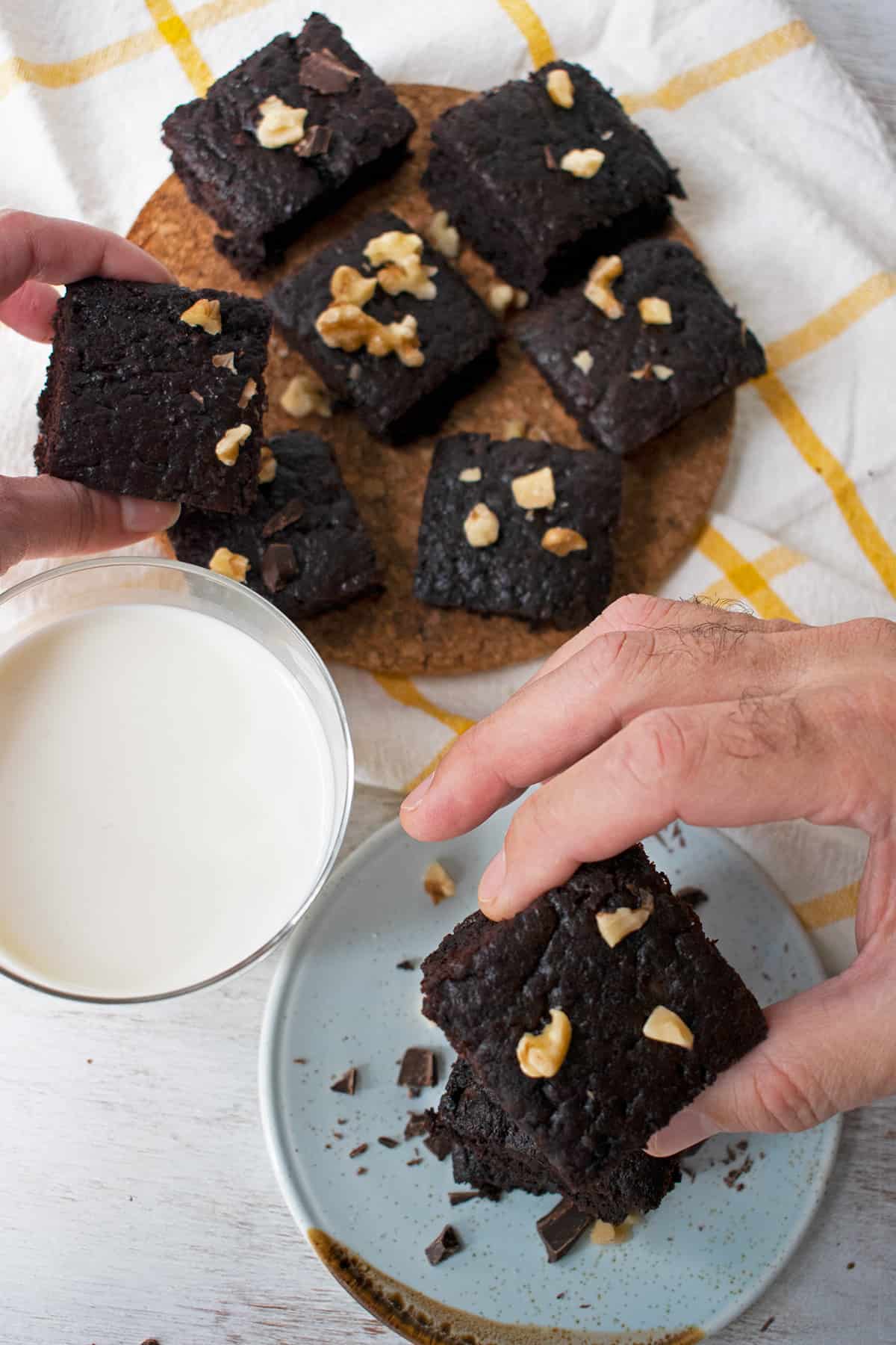 brownies sobre una mesa con un vaso de leche, las manos de dos personas cogiendo algunos de los brownies.