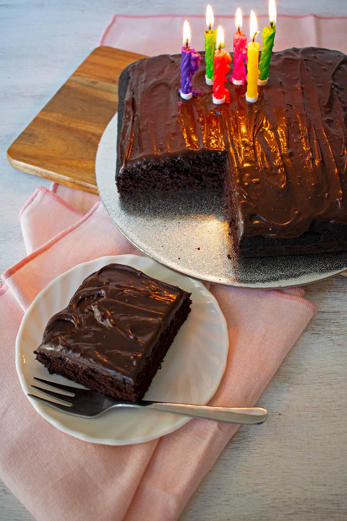 Pastel de chocolate con velas de cumpleaños de diferentes colores y una porción de torta en un plato blanco con un tenedor aparte.