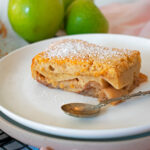 tarta de manzana casera servida en un plato blanco con una pequeña cuchara y tres manzanas verdes detrás