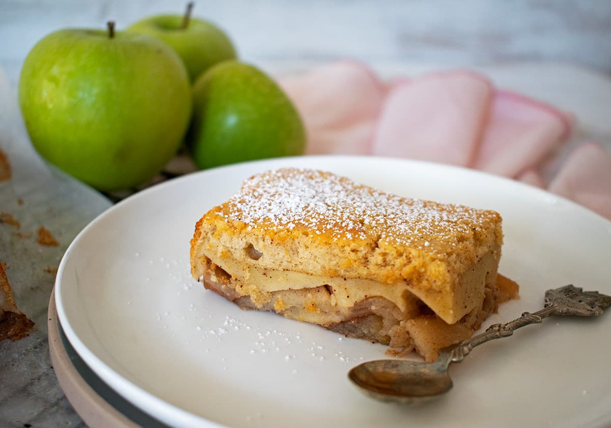 porción de torta mágica de manzana servida en un plato blanco acompañado de 3 manzanas verdes.