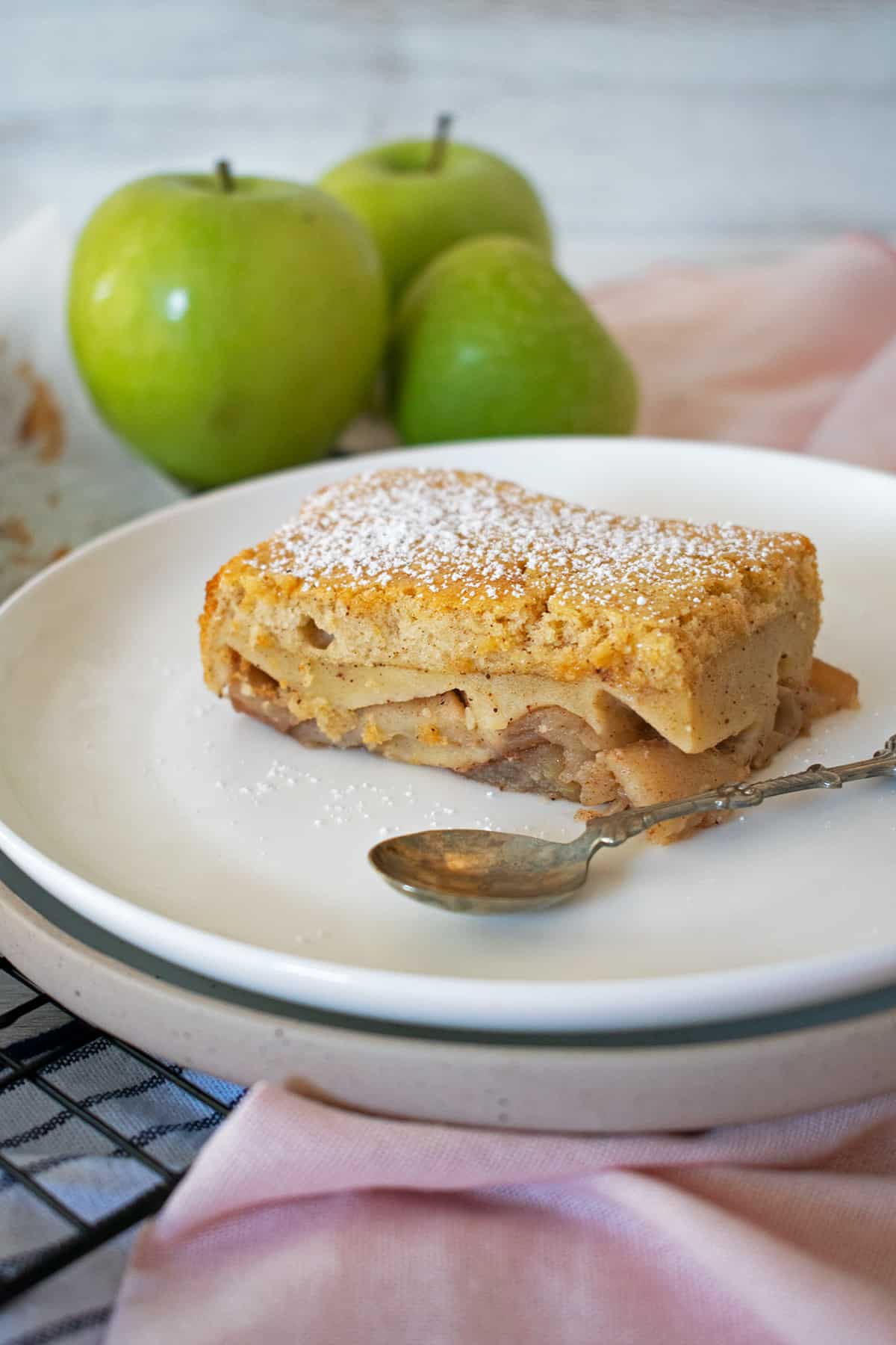 Tarta de manzana servida en un plato blanco con una pequeña cuchara, tres manzanas verdes detrás.