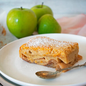 tarta de manzana casera servida en un plato blanco con tres manzanas verdes atrás.