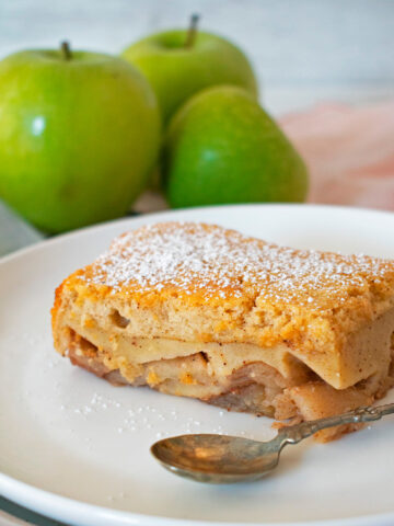 tarta de manzana casera servida en un plato blanco con tres manzanas verdes atrás.