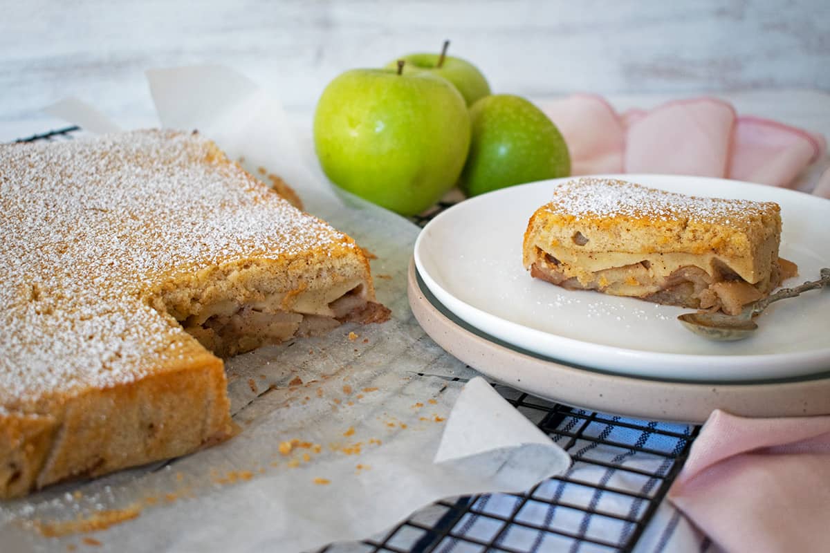 Tarta de manzana sobre una rejilla negra, una porción servida en un plato con una pequeña cuchara y tres manzanas verdes atrás.