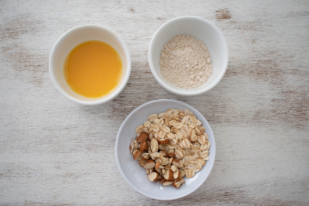 3 contenedores redondos blancos uno de ellos con harina de avena, otro con huevo batido y otro con almendras y avena en hojuelas