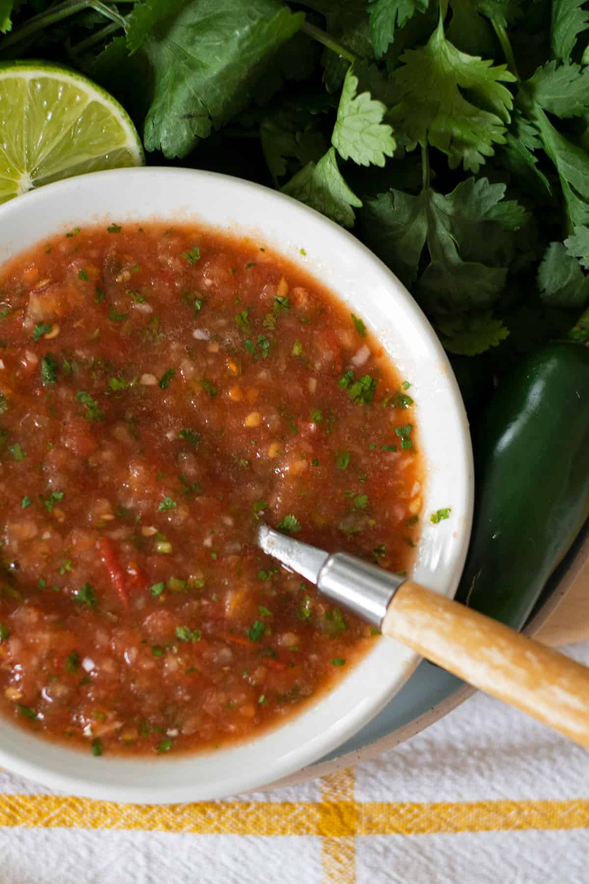 salsa fresca estilo restaurante servida en un plato blanco con una cuchara de madera un manojo de cilantro, limón y un jalapeño.