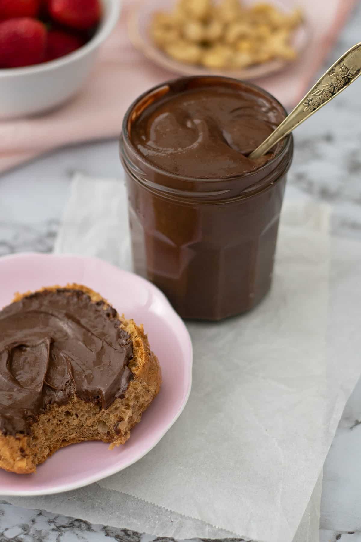 Nutella sin azúcar en un frasco de vidrio acompañada de un pan redondo sobre un plato rosado redondo