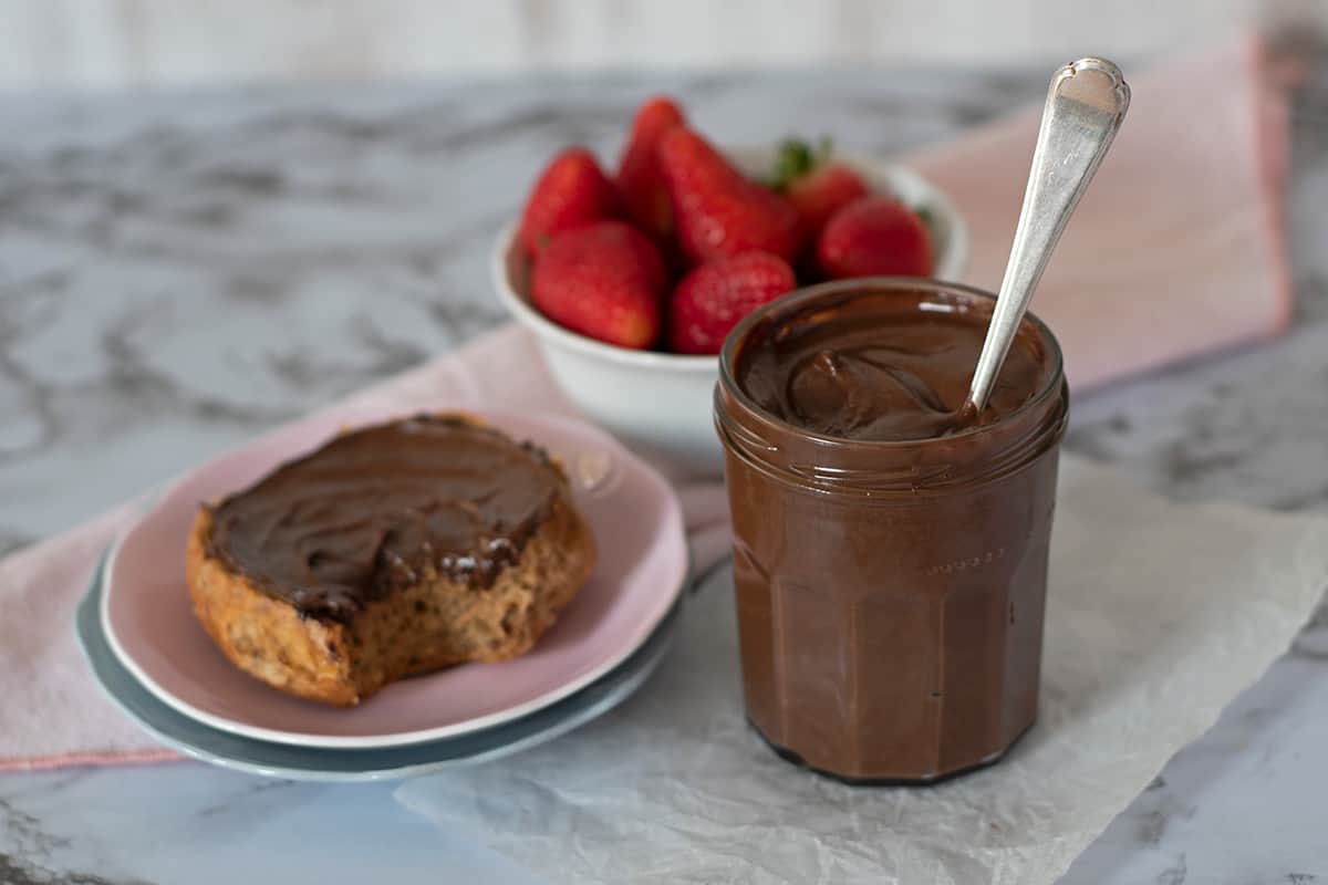 Un frasco de vidrio con crema de cacao casera acompañada de un pan y un bol de fresas