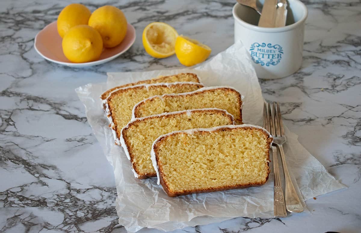 porciones de torta de limón servidas sobre una superficie de mármol acompañadas por dos tenedores plateados con tres limones en la parte de atrás servidos en un pequeño plato rosado.