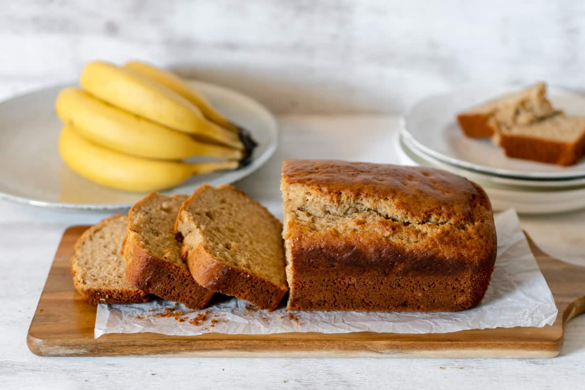 Banana bread casero partido en rebanados servido sobre una tabla de madera acompañando de bananos frescos.