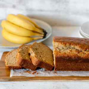 Pan de banana partido en rebanadas servido sobre una tabla de madera acompañado de bananos frescos