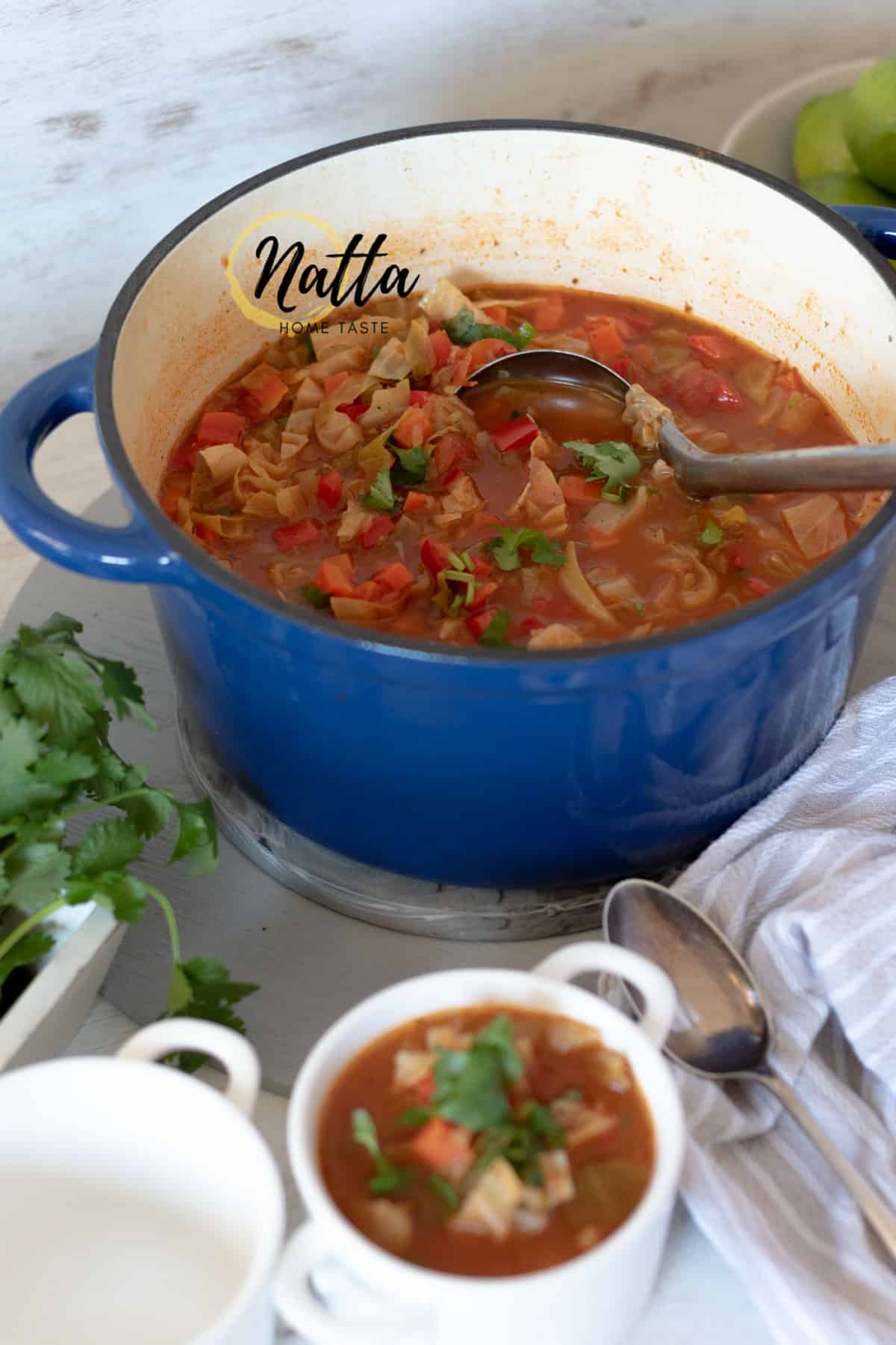 Sopa de repollo servida en una olla azul acompañada de recipientes blancos y cucharas