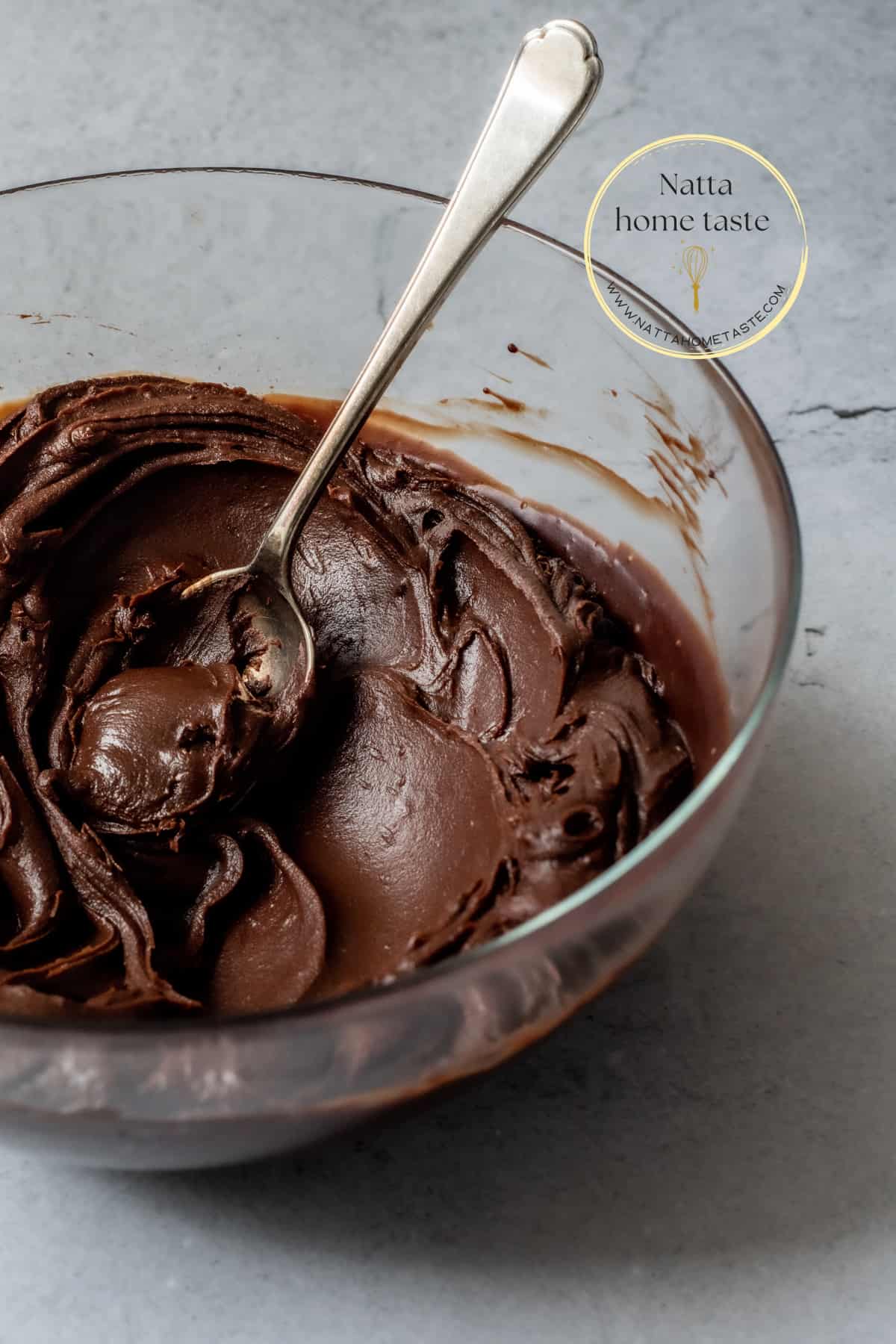 ganache de chocolate para cobertura en un bol transparente con una cuchara plateada sobre una mesa gris.