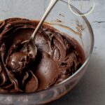 Ganache de chocolate para cobertura servido en un bol transparente con una cuchara plateada sobre una mesa gris.