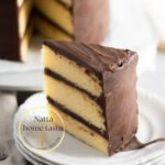 porción de torta de vainilla cubierta de ganache de chocolate servida en un plato blanco.
