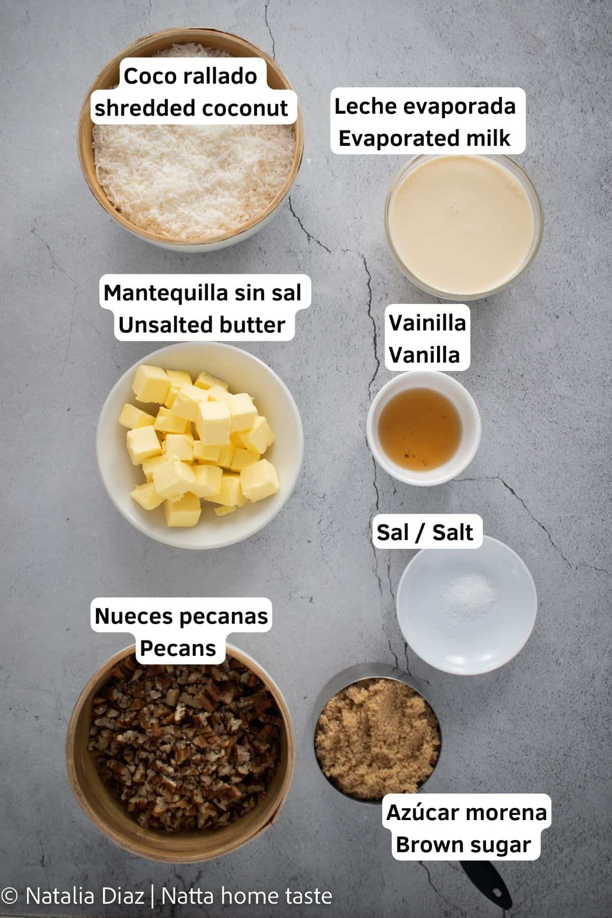 Ingredientes para preparar relleno o betún de coco y nueces. Los ingredientes están servidos en contenedores separados sobre una mesa de piedra gris. Coco rallado, leche evaporada, mantequilla sin sal, extracto de vainilla, nueces pecanas, azúcar morena y sal.