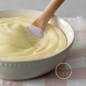 Glaseado de queso crema servido en un bol color crema con una espátula de silicona con mango de madera. el bol esta sobre una servilleta de tela con cuadros rosados y blancos.