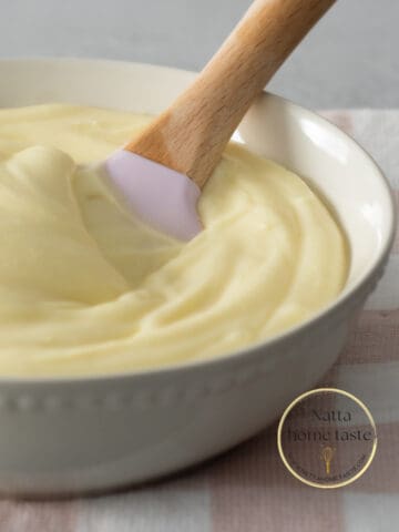 Glaseado de queso crema servido en un bol color crema con una espátula de silicona con mango de madera. el bol esta sobre una servilleta de tela con cuadros rosados y blancos.