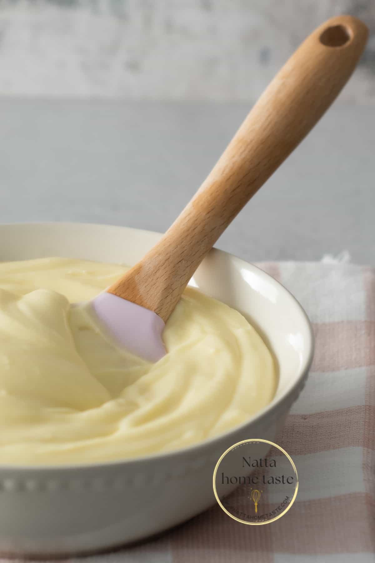 Glaseado de queso crema servido en un bol color crema con una espátula de silicona con mango de madera. el bol esta sobre una servilleta de tela con cuadros rosados y blancos. 