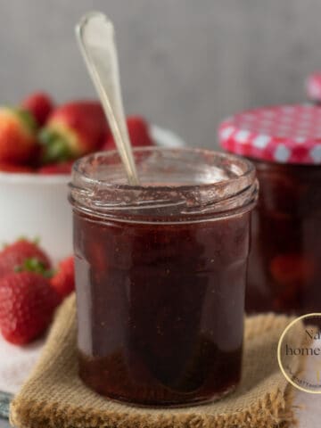 frasco de vidrio con mermelada de fresa casera y una cuchara dentro del frasco. Fresas frescas en la parte de atrás.