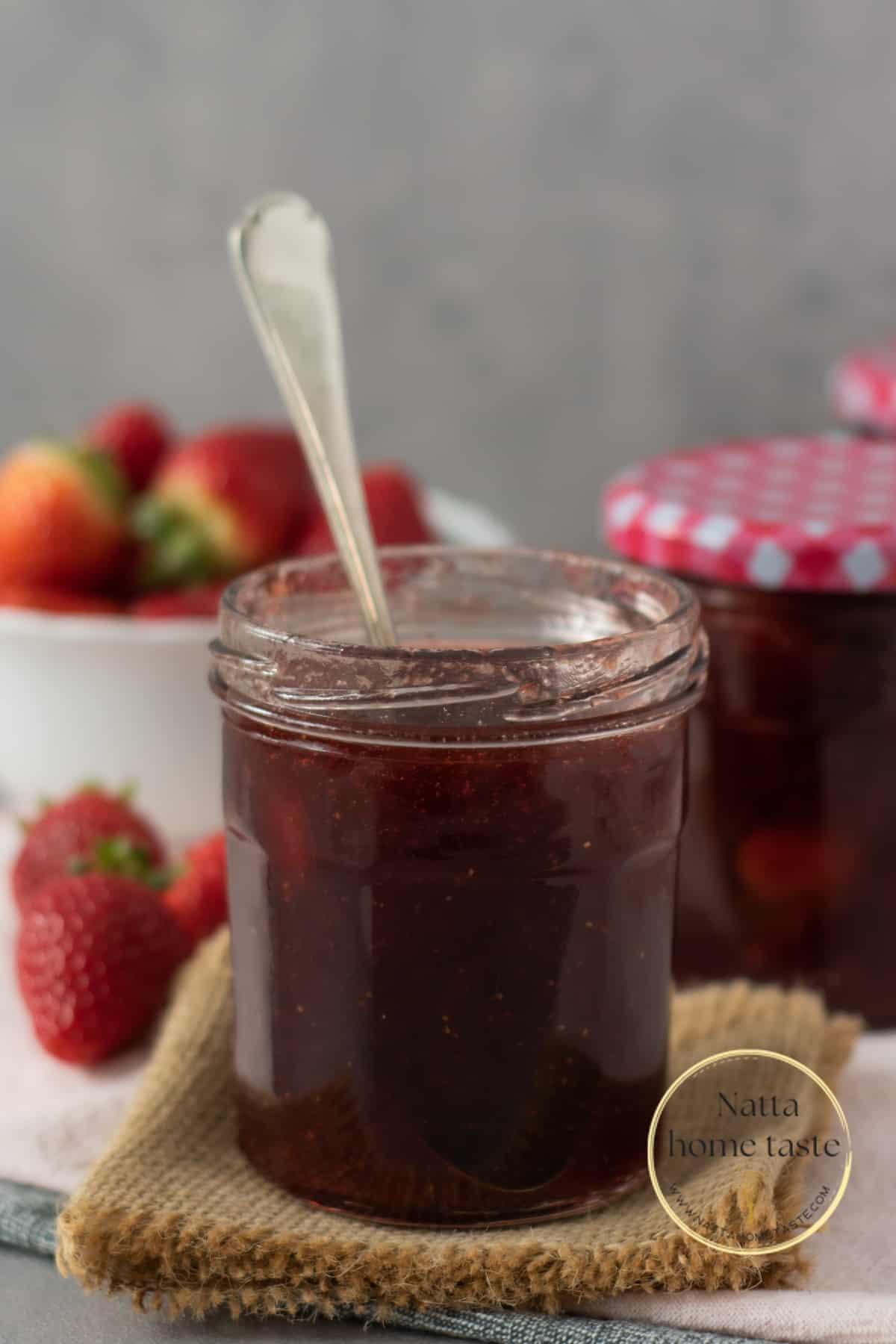 frasco de vidrio con mermelada de fresa casera y una cuchara dentro del frasco. Fresas frescas en la parte de atrás.