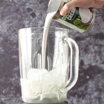 Vaso de una licuadora con cubos de hielo y alguien vertiendo coconut cream al vaso. 