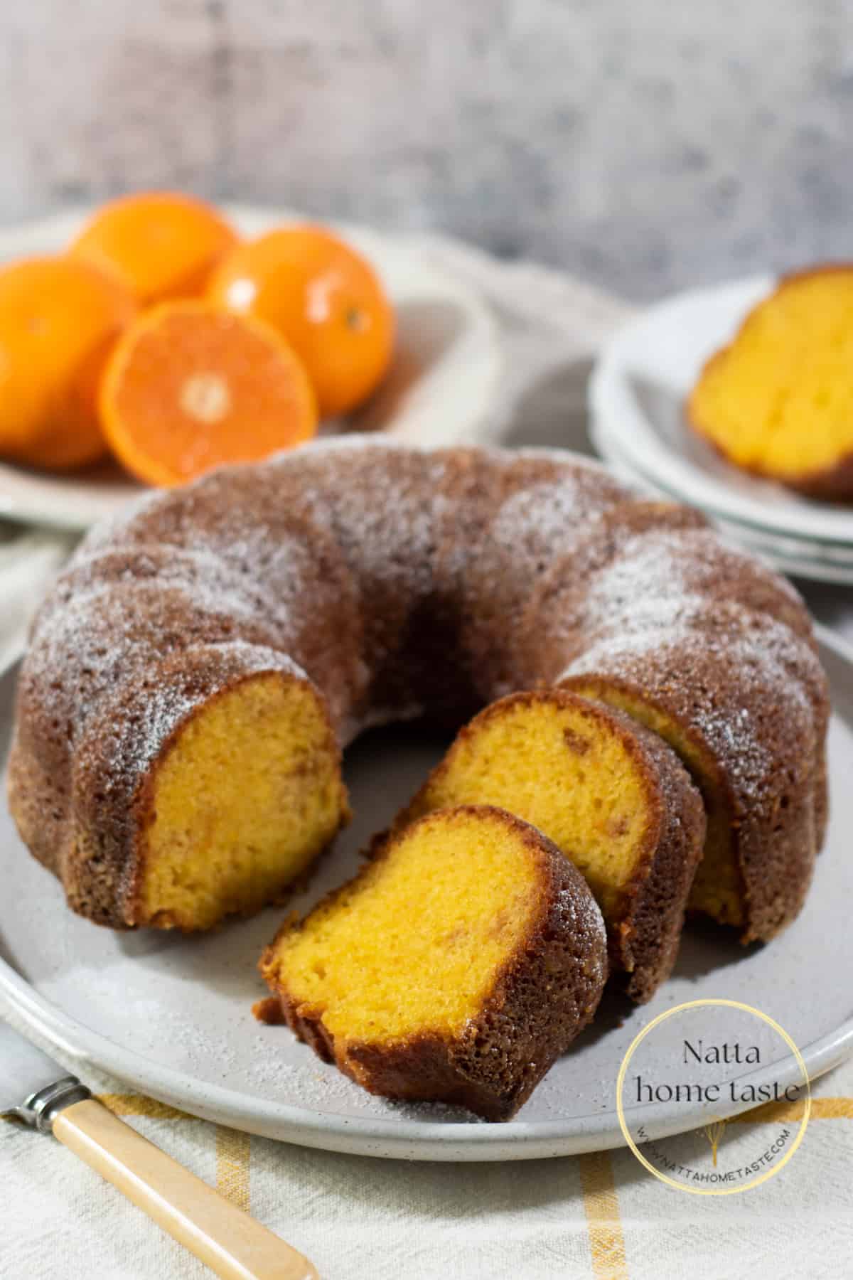 Torta de mandarina redonda servida en un plato blanco con mandarinas frescas en la parte de atrás.