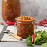 frasco de vidrio con ají picante colombiano sobre una mesa con una pequeña cuchara al lado y un manojo de cilantro fresco.