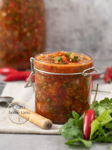 frasco de vidrio con ají picante colombiano sobre una mesa con una pequeña cuchara al lado y un manojo de cilantro fresco.