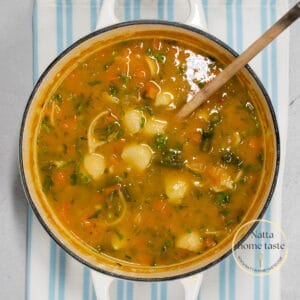 imagen desde arriba de una olla blanca con sopa de pasta con verduras y una cuchara de madera.