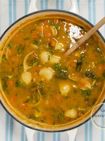 imagen desde arriba de una olla blanca con sopa de pasta con verduras y una cuchara de madera.