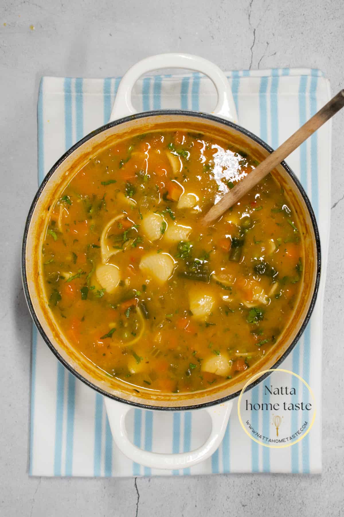 imagen desde arriba de una olla blanca con sopa de pasta con verduras y una cuchara de madera.