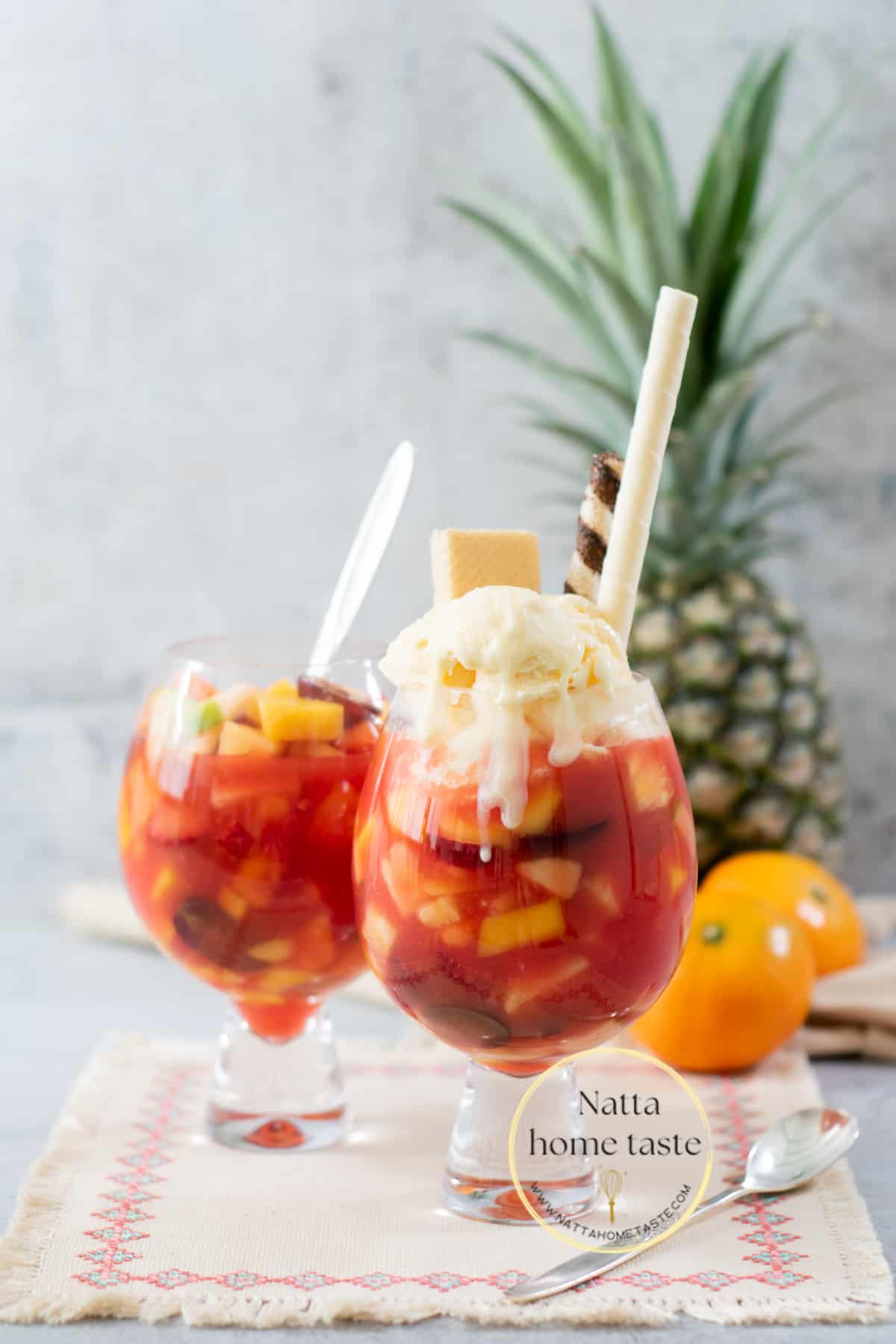 dos copas de salpicón de frutas colombino con helado de vainilla y galletas wafer con una piña entera y los mandarinas en la parte de atrás.