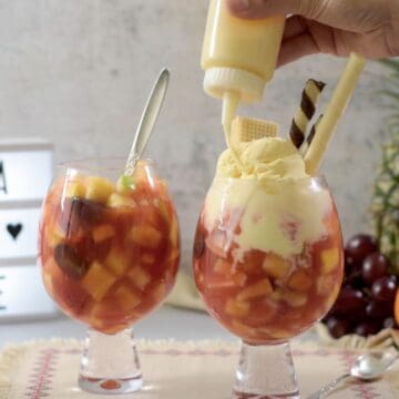 dos copas de salpicón de frutas colombino con helado de vainilla y galletas wafer con una piña entera y los mandarinas en la parte de atrás.