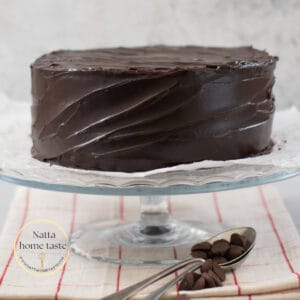 torta de chocolate cubierta con ganache de chocolate sobre una base de cristal sobre una mesa con un mantel beige con rayas rojas. Dos cucharas metálicas con trozos de chocolate al lado de la base que sostiene el pastel.