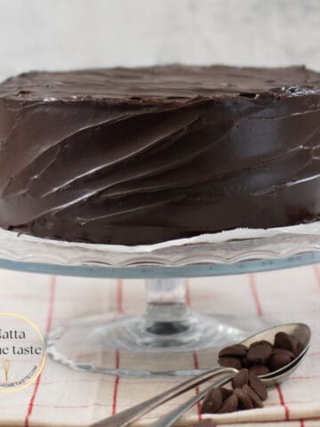 torta de chocolate cubierta con ganache de chocolate sobre una base de cristal sobre una mesa con un mantel beige con rayas rojas. Dos cucharas metálicas con trozos de chocolate al lado de la base que sostiene el pastel.