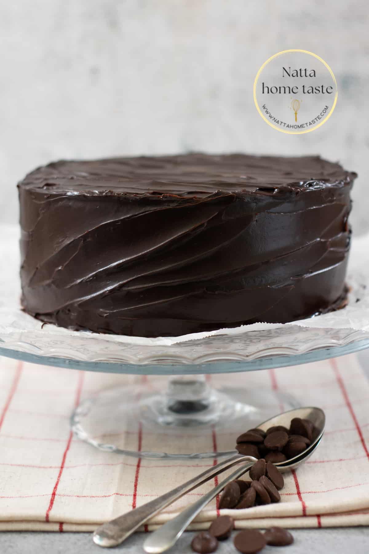 torta de chocolate húmeda cubierta con ganache de chocolate sobre una base de cristal sobre una mesa con un mantel beige con rayas rojas. Dos cucharas metálicas con trozos de chocolate al lado de la base que sostiene el pastel.