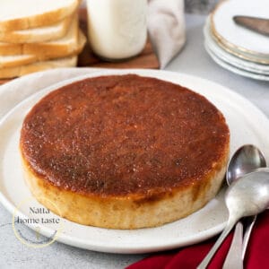 budín de pan redondo sobre un plato blanco con pan tajado y un vaso de leche en la parte de atrás.