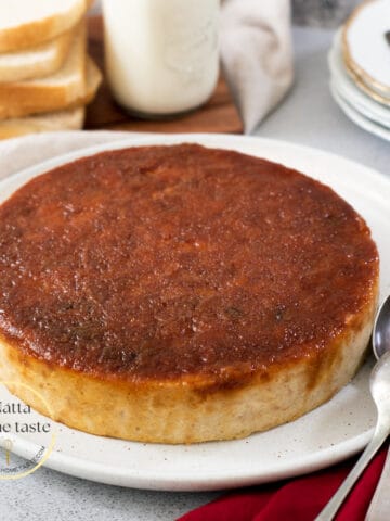 budín de pan redondo sobre un plato blanco con pan tajado y un vaso de leche en la parte de atrás.