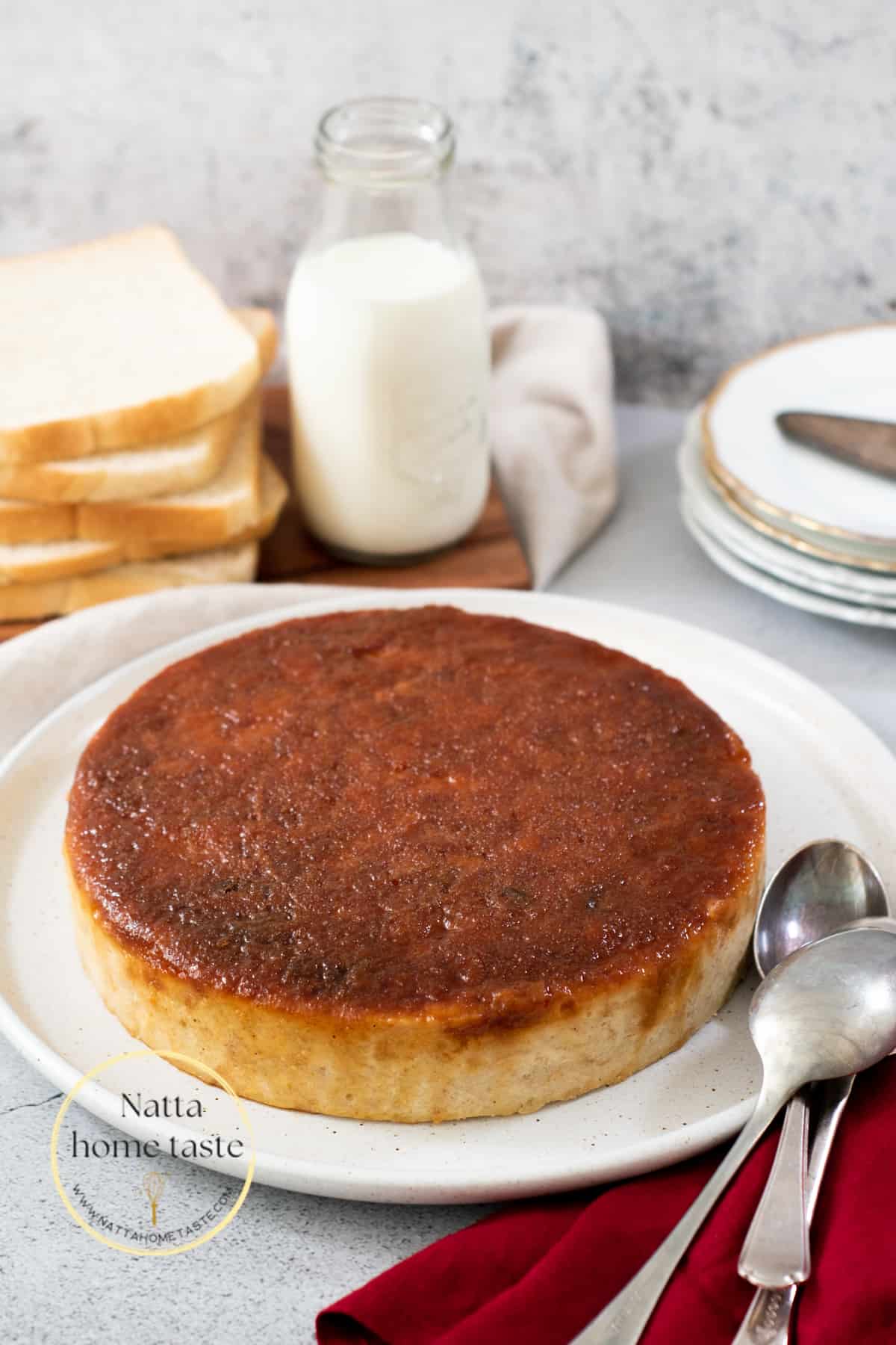 budín de pan redondo sobre un plato blanco con pan tajado y un vaso de leche en la parte de atrás.