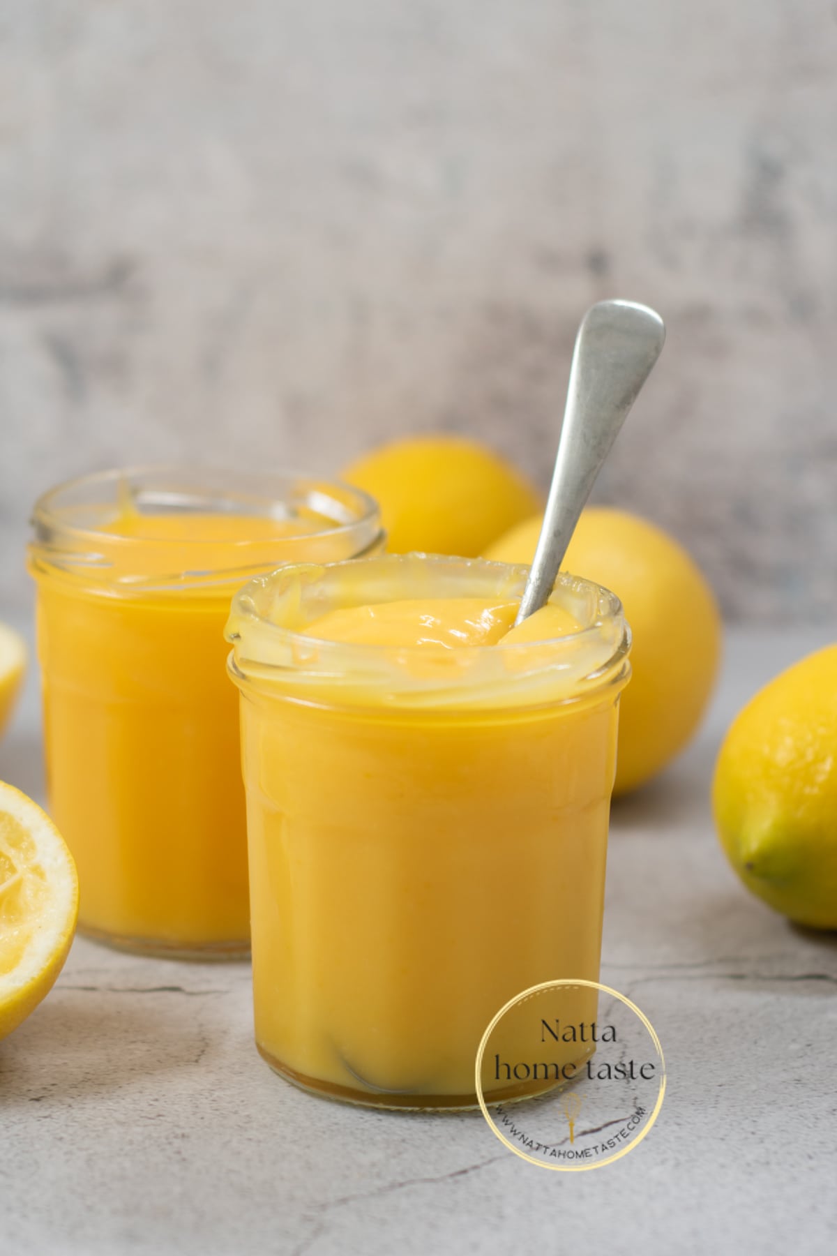 crema de limón servida en dos frascos de vidrio con una cuchara y varios limones amarillos alrededor.
