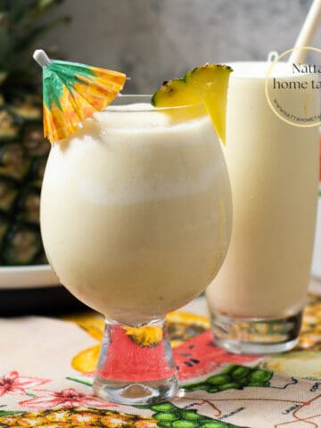 dos vasos con piña colada, un vaso con una sombrilla de coctel y el otro con un pitillo de madera. Una piña entera en la parte de atrás.