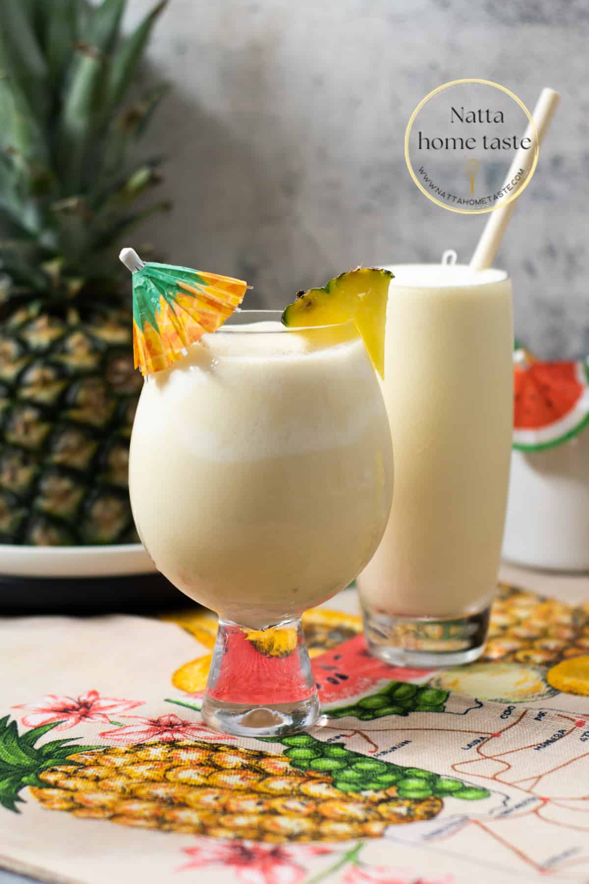 dos vasos con piña colada, un vaso con una sombrilla de coctel y el otro con un pitillo de madera. Una piña entera en la parte de atrás. 