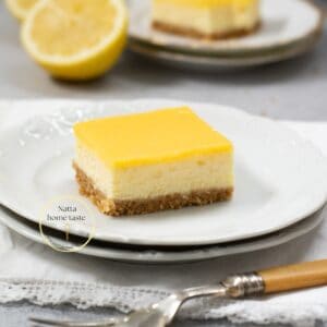 cheesecake de limón partido en una porción cuadrada sobre un plato blanco redondo con medio limón en la parte de atrás
