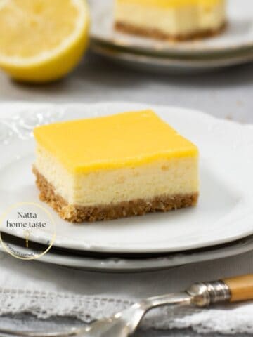 cheesecake de limón partido en una porción cuadrada sobre un plato blanco redondo con medio limón en la parte de atrás