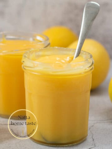 crema de limón servida en dos frascos de vidrio con una cuchara y varios limones amarillos alrededor.