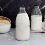 leche de avena casera en un frasco de vidrio sobre una mesa con una taza de avena en copos en la parte de atrás.